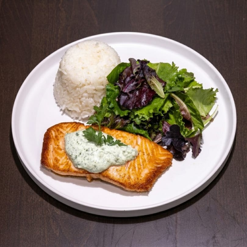 A salmon fillet with green sauce, white rice, and a side salad on a white plate.