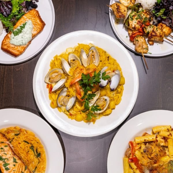 Four plates of food, including salmon, paella with seafood, chicken skewers, and pasta.