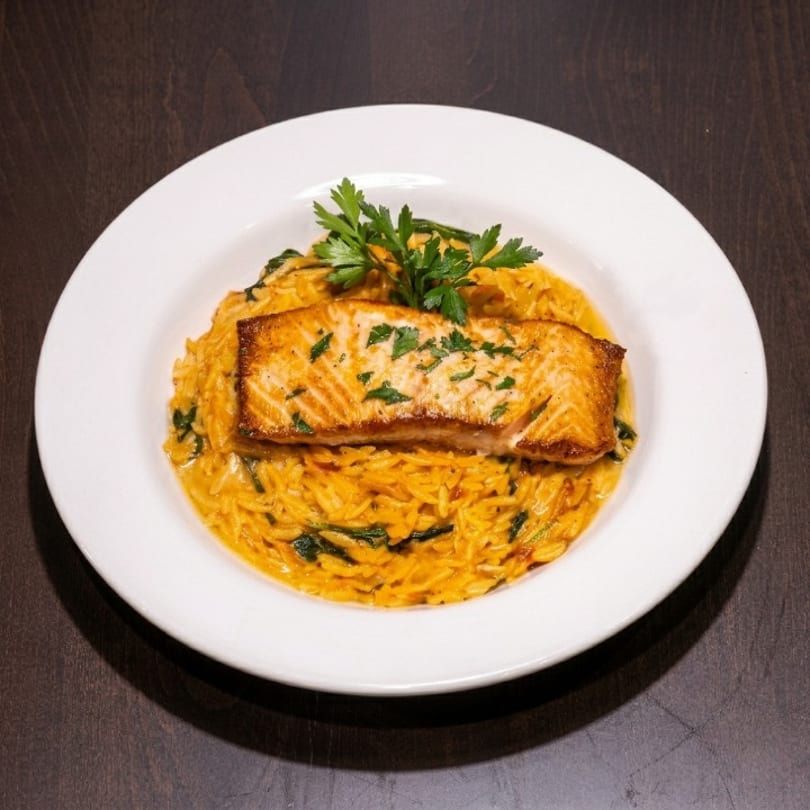 Pan-seared salmon fillet over creamy orange orzo pasta with spinach, garnished with parsley.